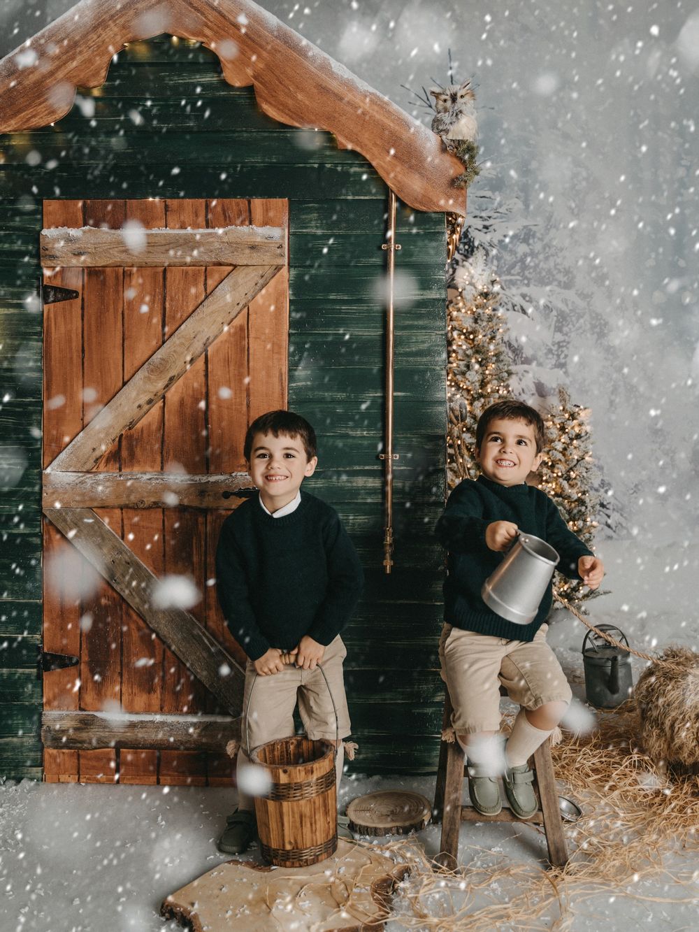Two children pose in front of a rustic barn door during a snowy winter photoshoot with Christmas decorations.