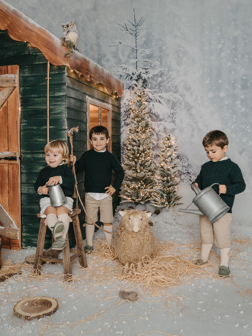 Three children holding watering cans by a rustic cabin with Christmas decorations.