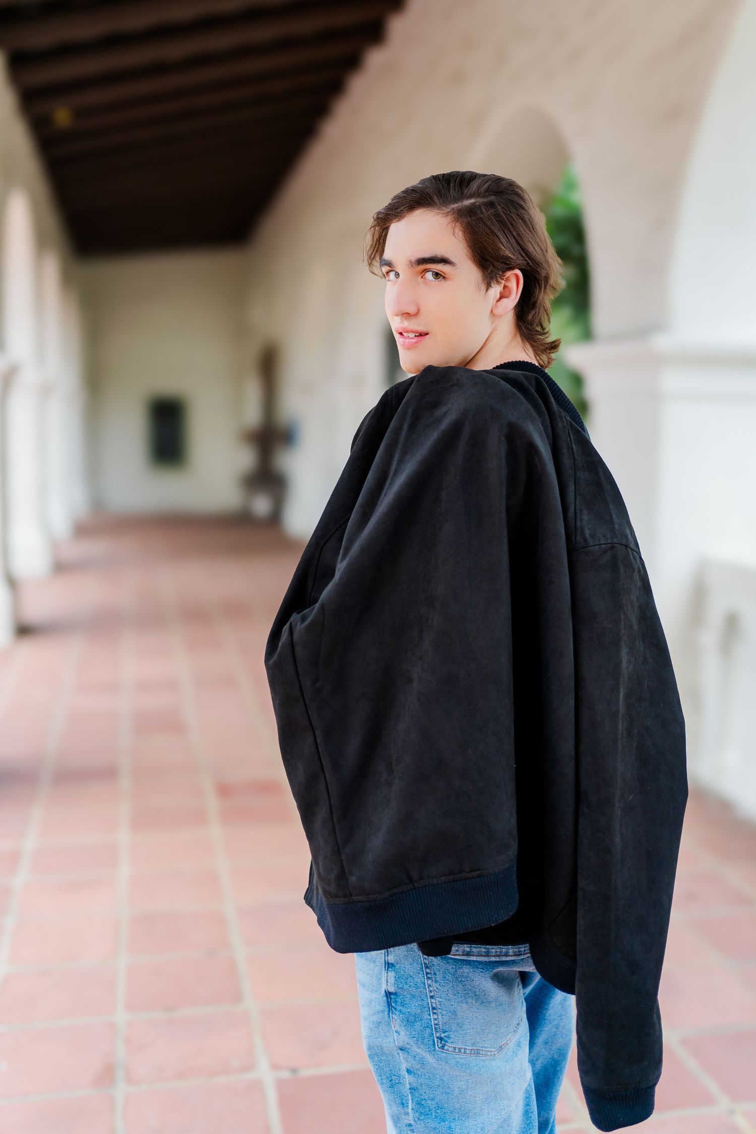 Person wearing a black oversized hoodie and blue jeans poses in profile against a Spanish-style corridor with arches.