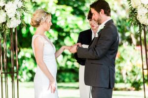 Wedding ceremony under a floral arch with couple exchanging rings in an outdoor garden setting.