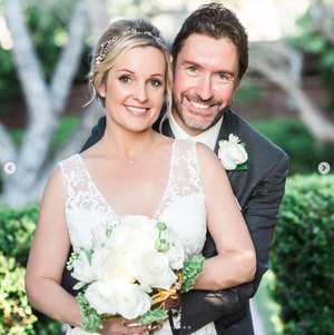 A wedding couple poses outdoors with white flowers and greenery in a garden setting.