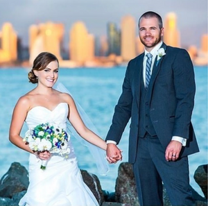 A wedding couple poses together on rocks by the water with a cityscape glowing in the sunset background.