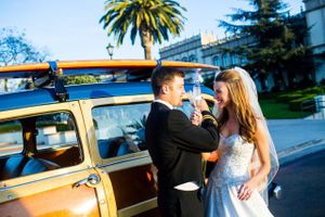 Couple in wedding attire poses beside vintage yellow car with surfboard on sunny day.