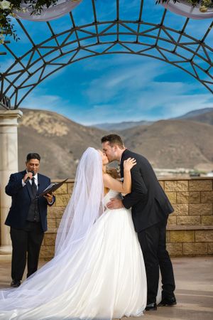 Mountain view wedding ceremony with couple kissing under architectural arch.
