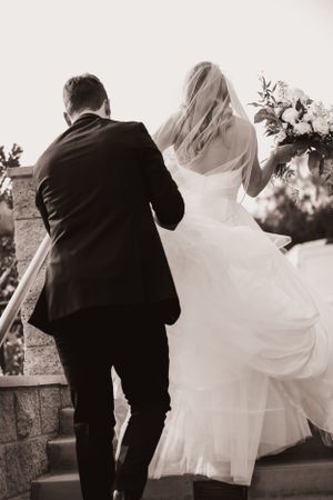 Black and white photo of couple walking up stairs in classic wedding attire.