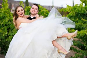 A joyful wedding photo at a vineyard showing a groom carrying his bride in a flowing white dress.
