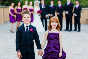 Ring bearer in navy suit and flower girl in purple dress hold hands at formal wedding.
