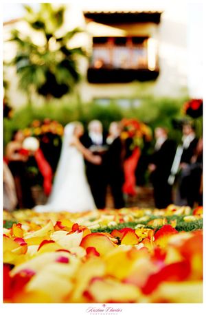 Rose petals scattered in foreground with wedding ceremony blurred in background.