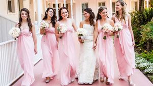 Bridesmaids in flowing pink dresses walking with a bride on a columned porch.