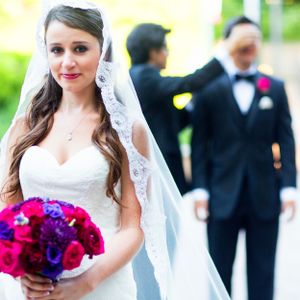 Bride in lace veil holds bright pink and purple bouquet with groom in background.