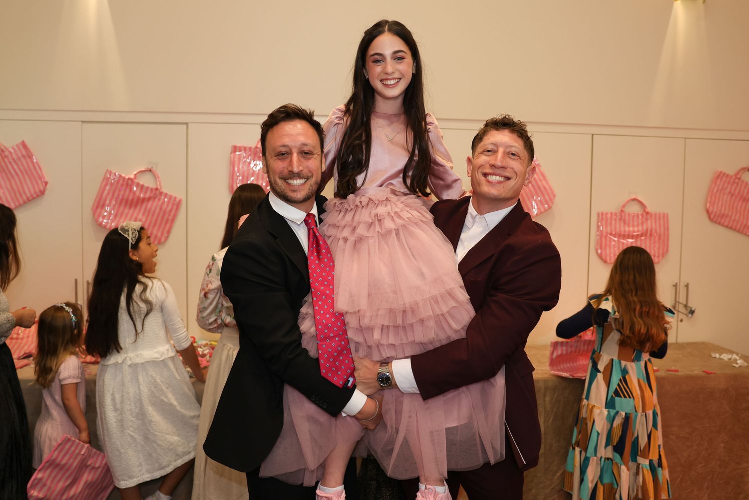 Young girl in pink tutu dress lifted by two smiling men in suits at Bat Mitzvah party.