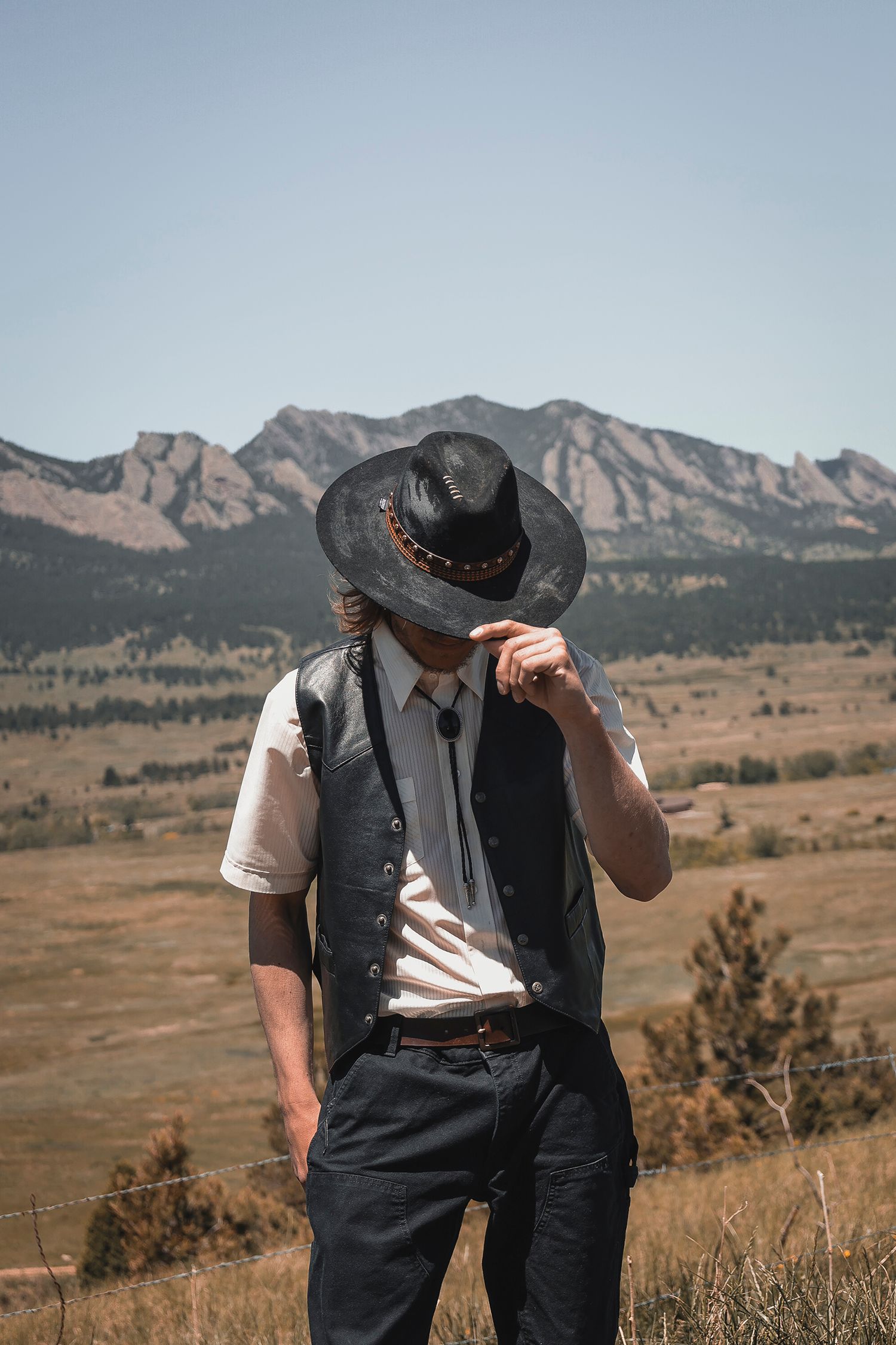 Stylishly dressed person in western attire with black hat and vest poses against scenic mountain landscape in Colorado.