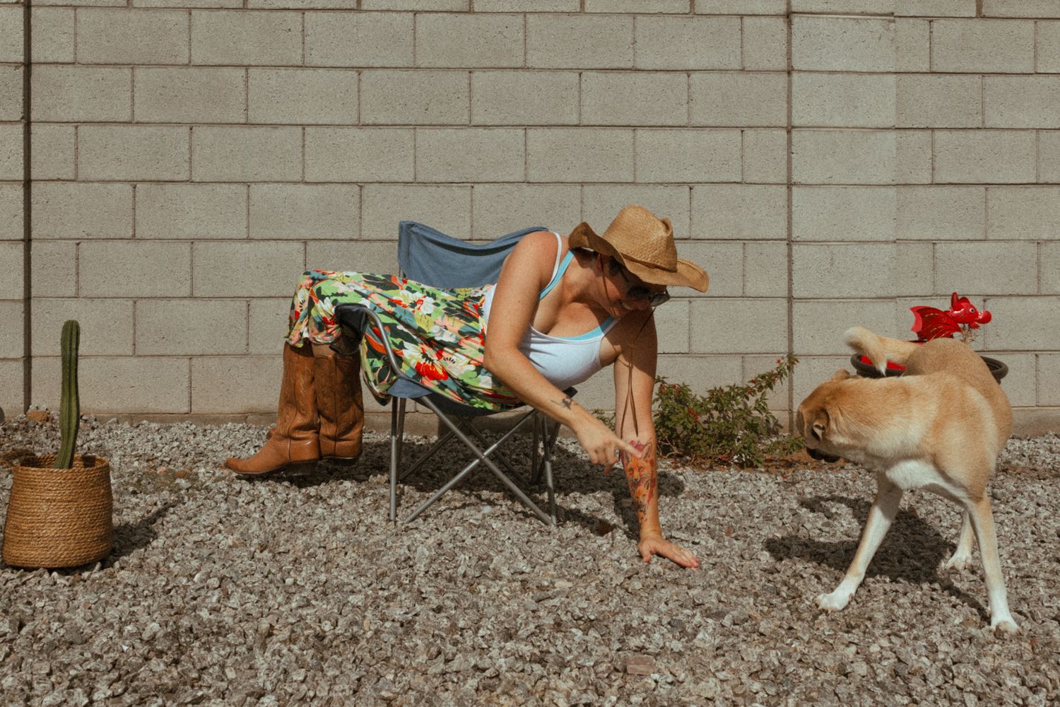 Woman in floral skirt and cowboy hat sits in camp chair, petting a dog near a cactus against a concrete wall. Whitney Popp, Phoenix Arizona