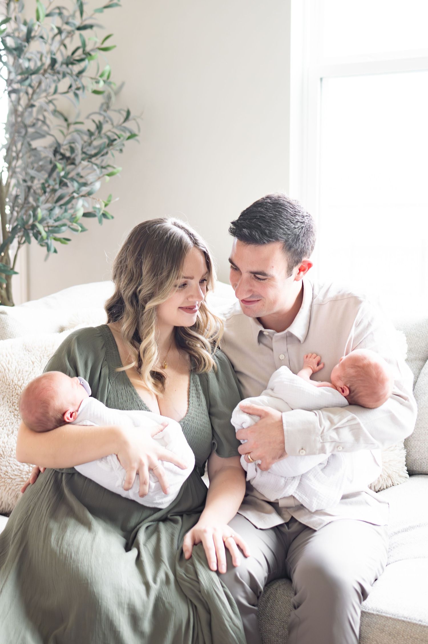 mom and dad stare lovingly into their twin boys arms.  one baby in each parents arms, while they sit on couch in living