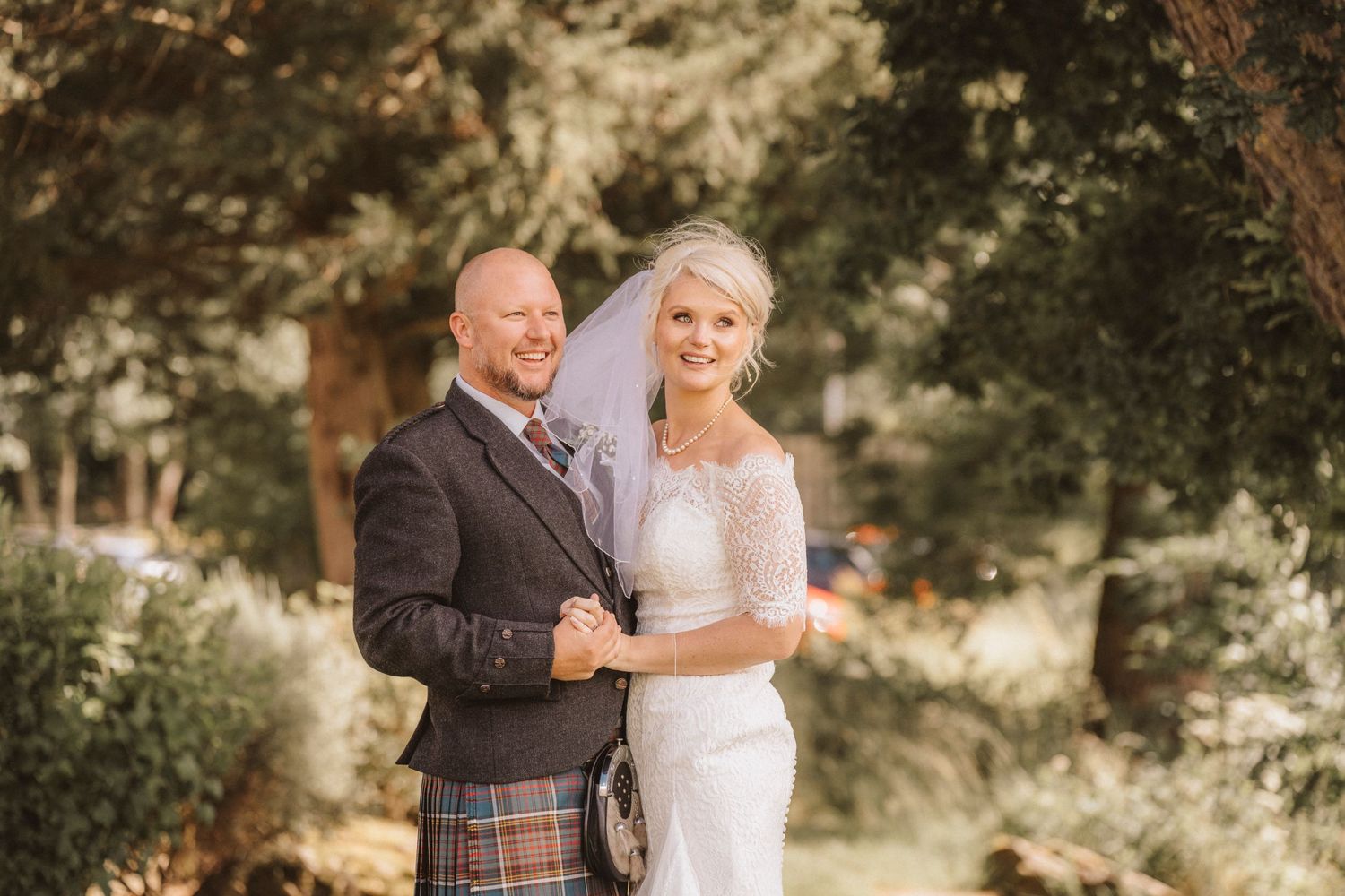 Wedding portrait featuring Scottish formal wear and elegant lace bridal gown in garden setting.