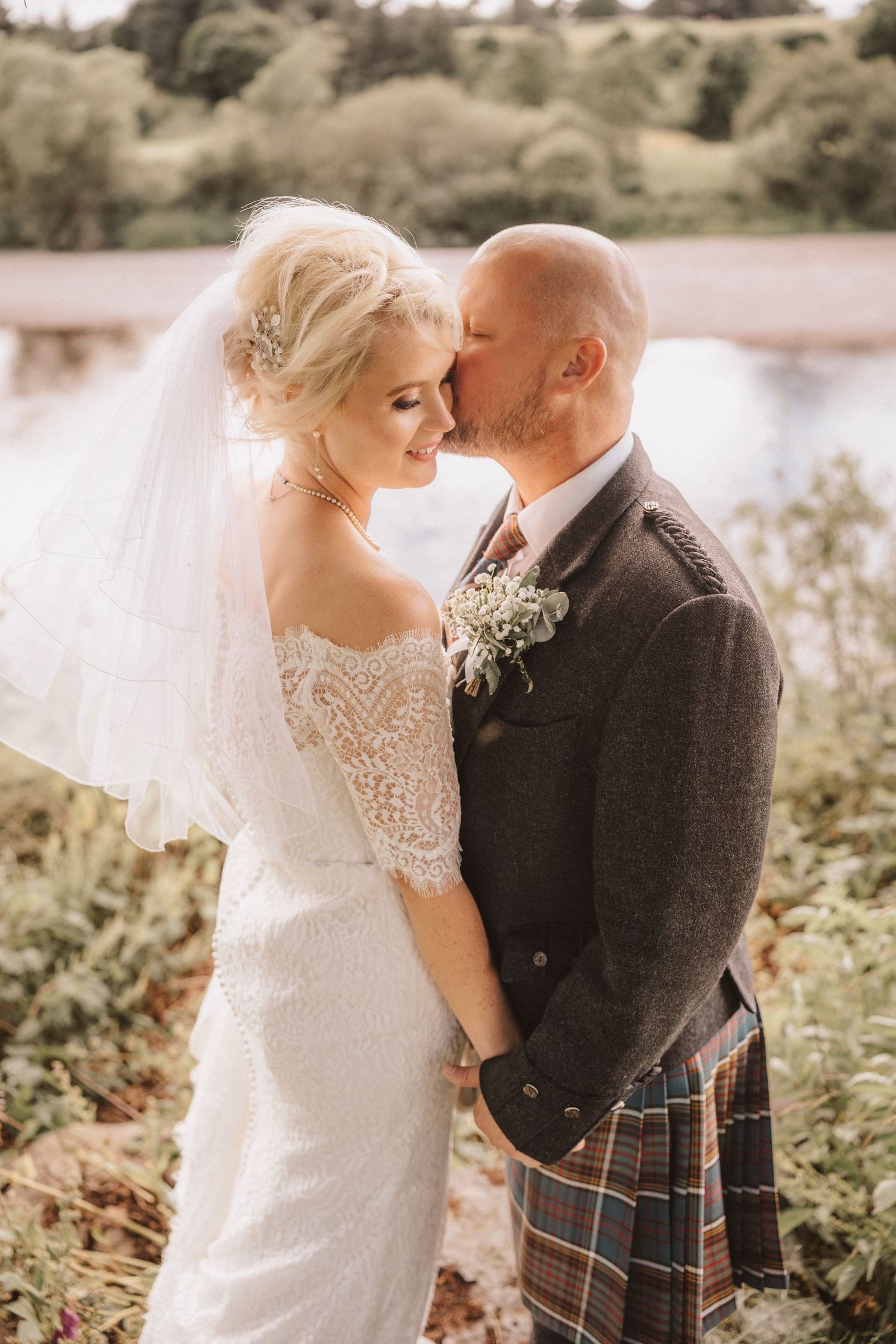 Intimate wedding moment captured by lake with groom in traditional Scottish attire.