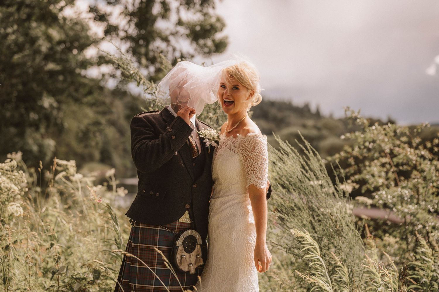 Joyful wedding portrait in Scottish highlands with traditional kilt and lace bridal gown.