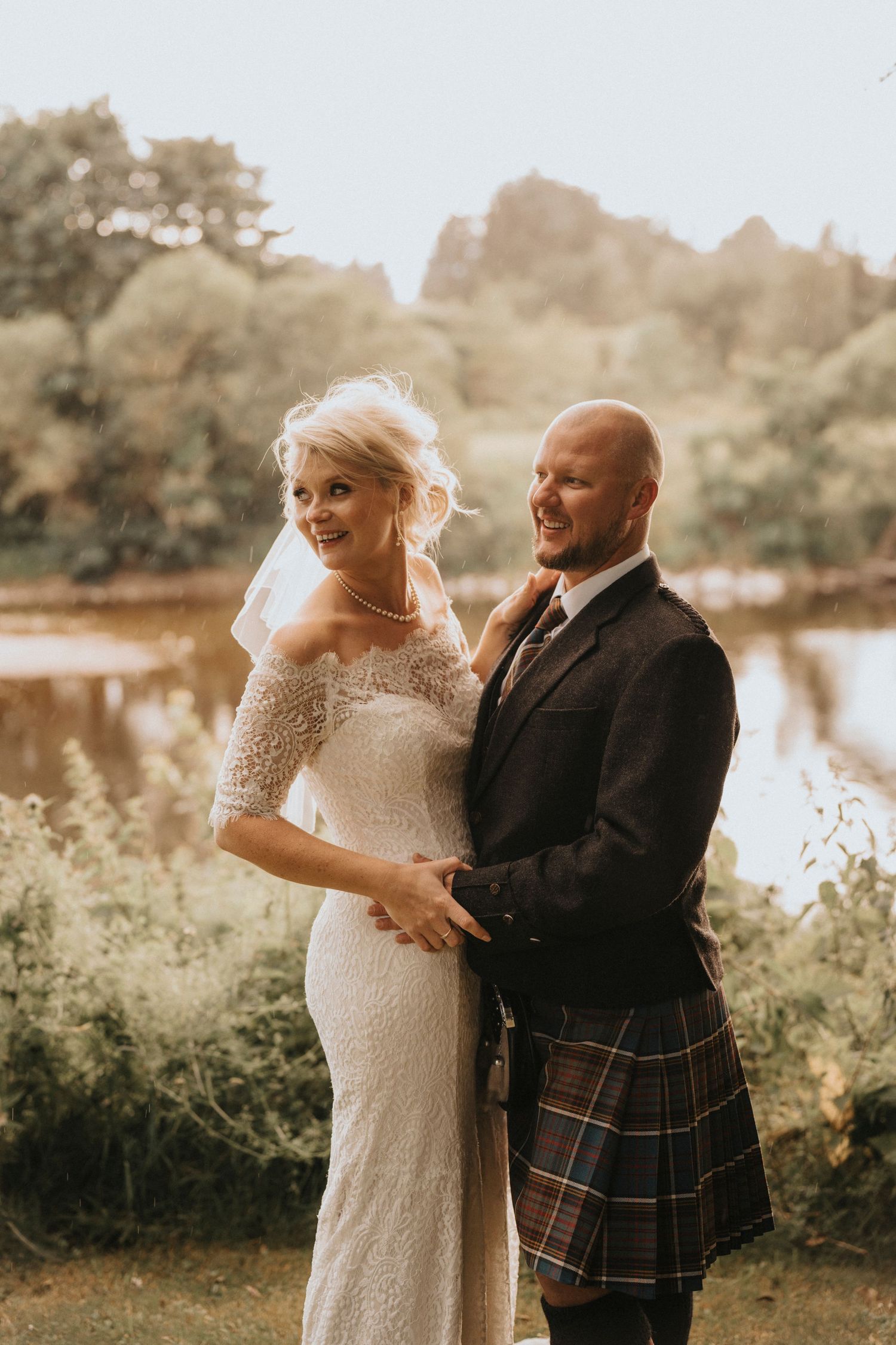 Scottish wedding couple sharing a tender moment by a lake with the groom in traditional kilt attire.