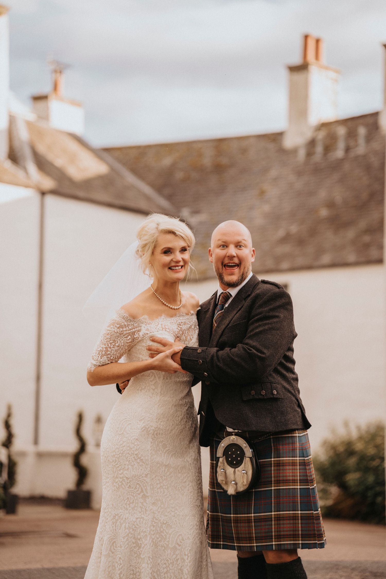 Happy newlyweds posing outside traditional white Scottish building with groom in traditional kilt.