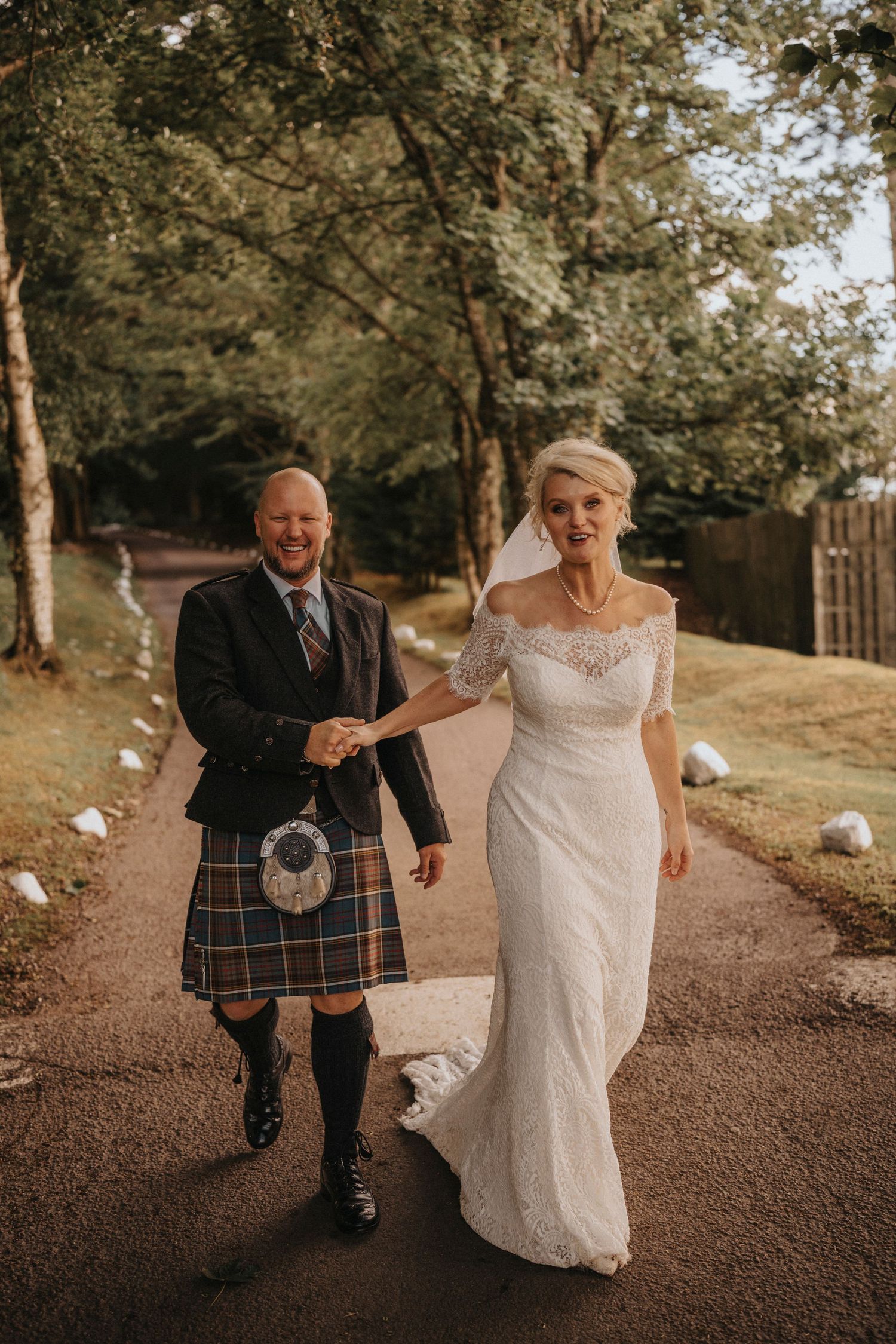 Candid shot of newlyweds walking along wooded path with groom in traditional Highland dress.