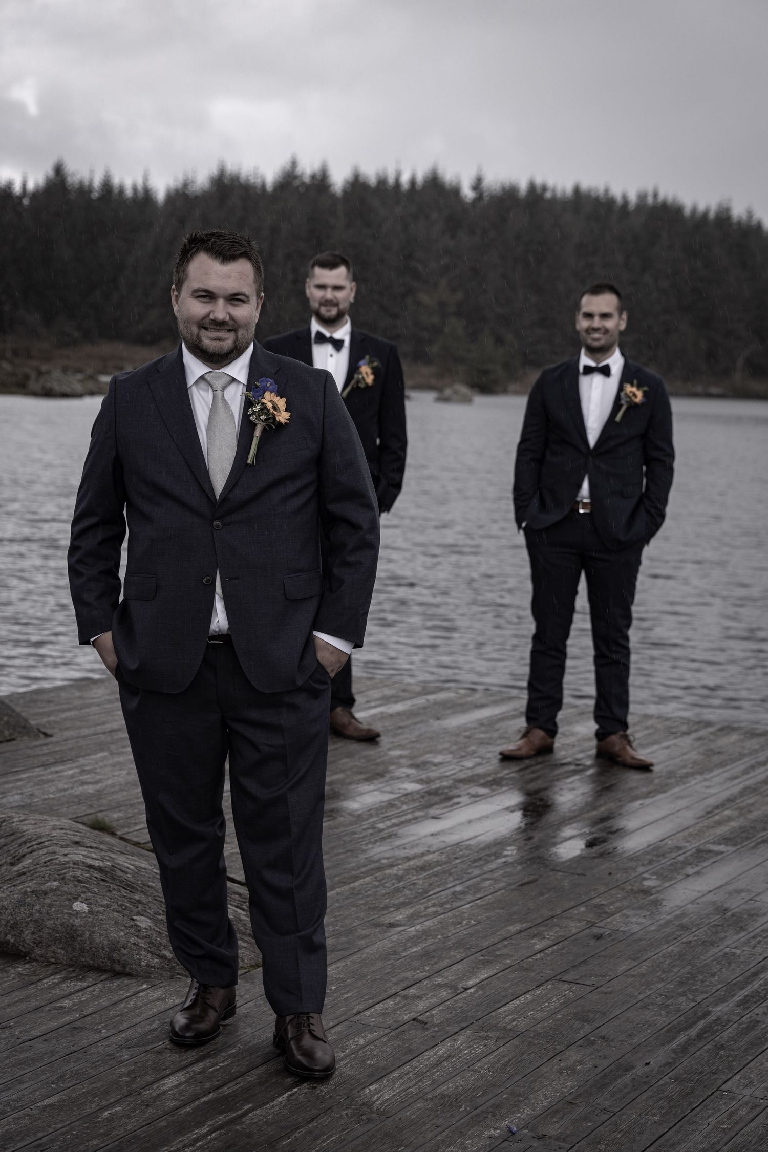 Groom with boutonniere stands with two groomsmen in dark suits on rainy waterfront dock.