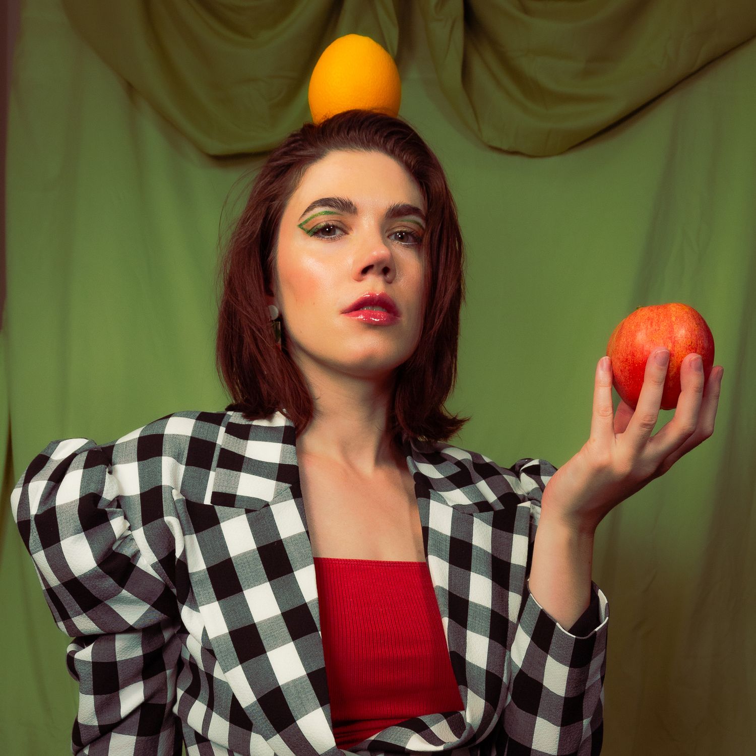 Artistic portrait of a woman in checkered jacket holding oranges against green backdrop.