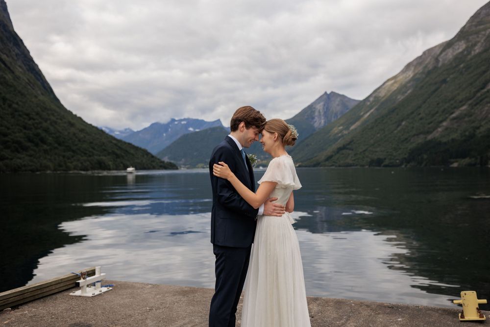 Newlyweds embrace on dock with mountains and lake creating stunning backdrop.