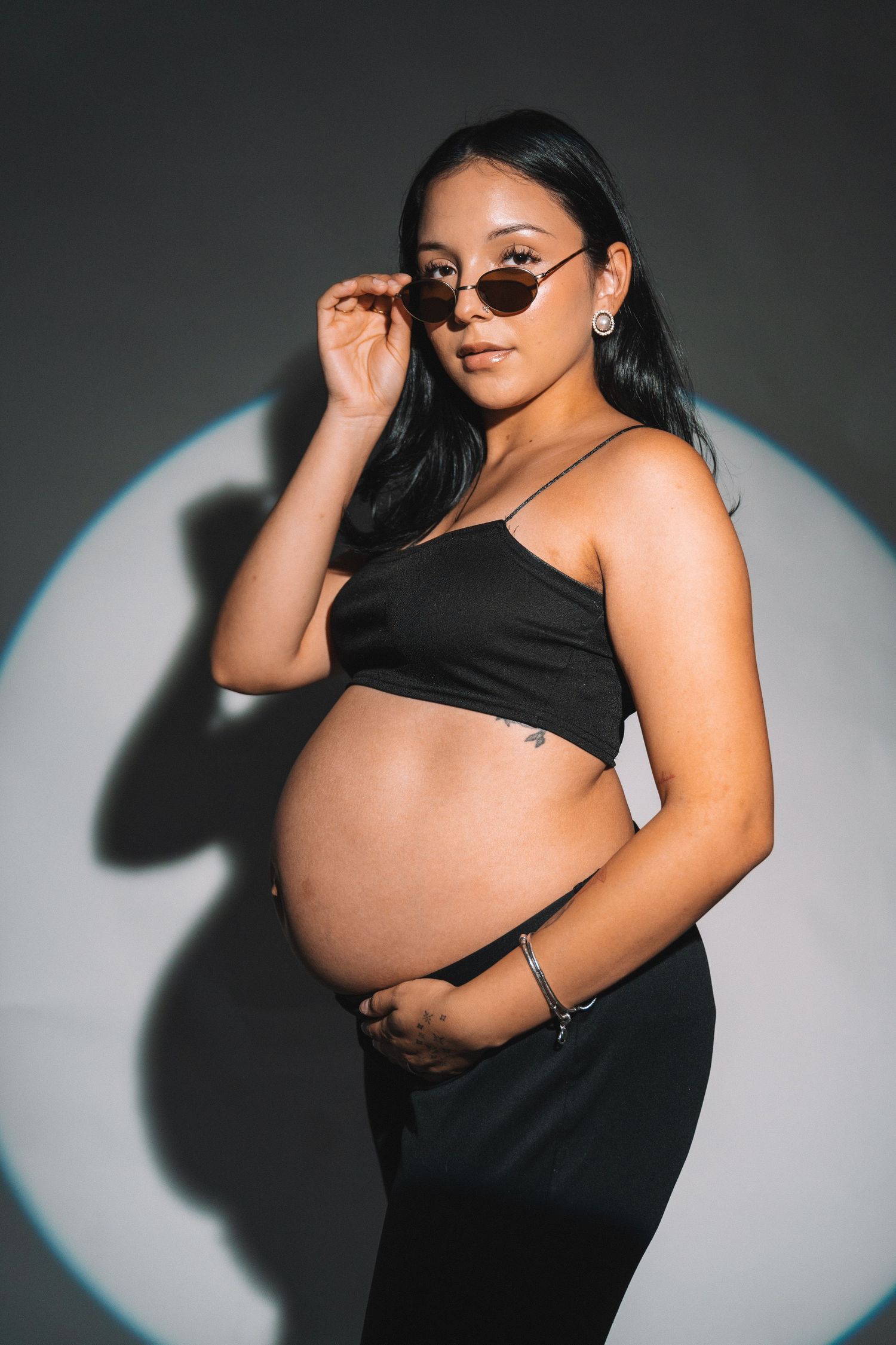 A stylish maternity photo of an expectant mother in black against dramatic lighting with sunglasses.