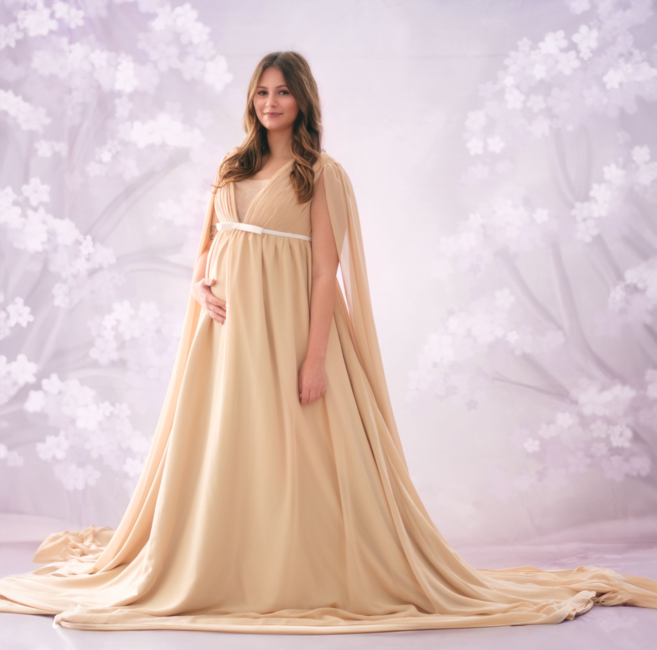 Elegant maternity portrait in flowing cream empire waist dress with cape against floral backdrop.