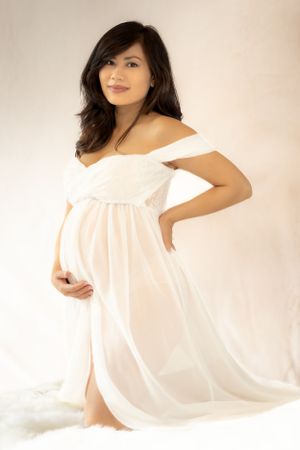 Elegant maternity portrait featuring white off-shoulder dress against soft beige background.