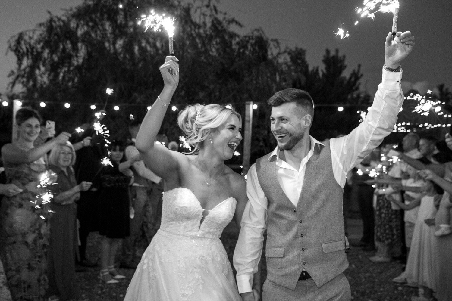 A newlywed couple holds sparklers and smiles joyfully while walking through a crowd of guests at an outdoor wedding
