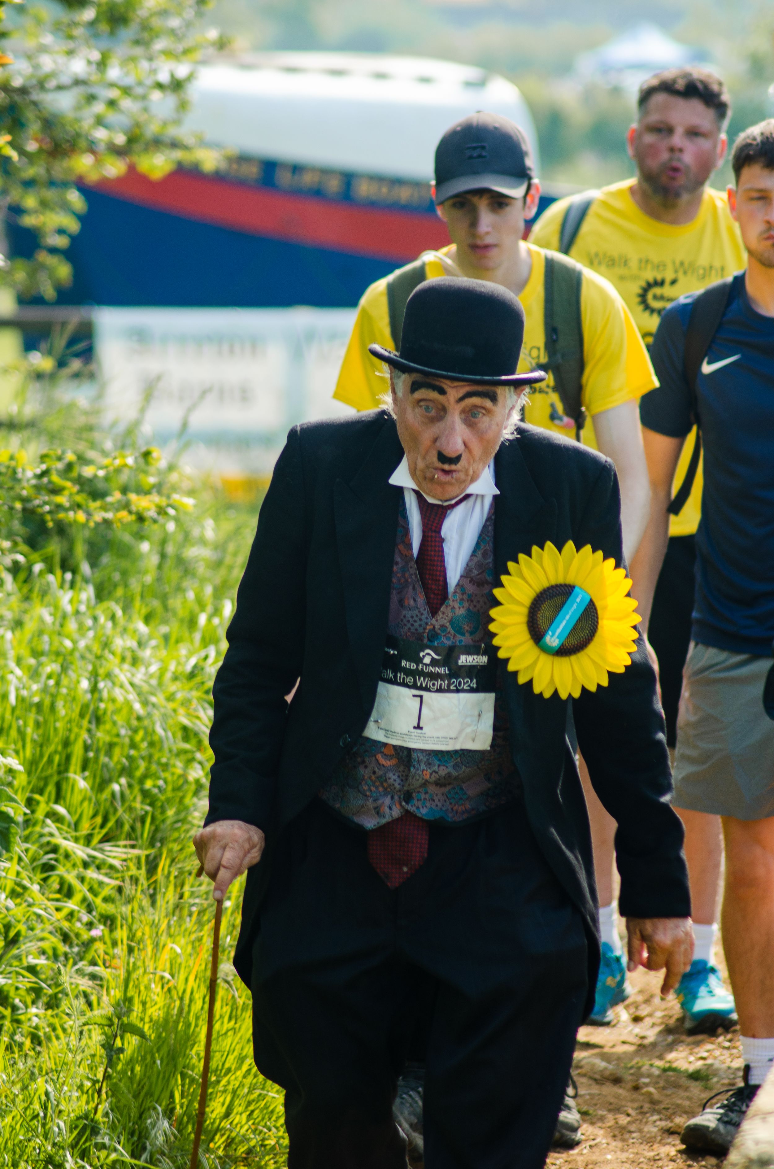 Person in Charlie Chaplin costume with sunflower pin participating in outdoor event.