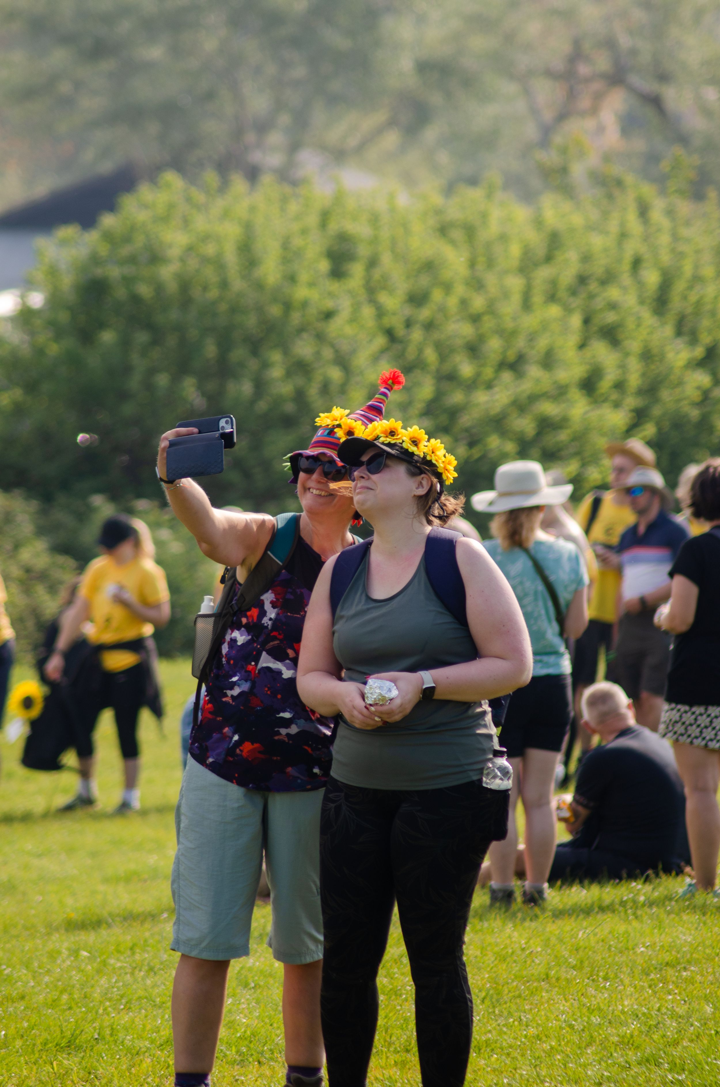 Event participants wearing sunflower crowns taking selfies on sunny hillside.