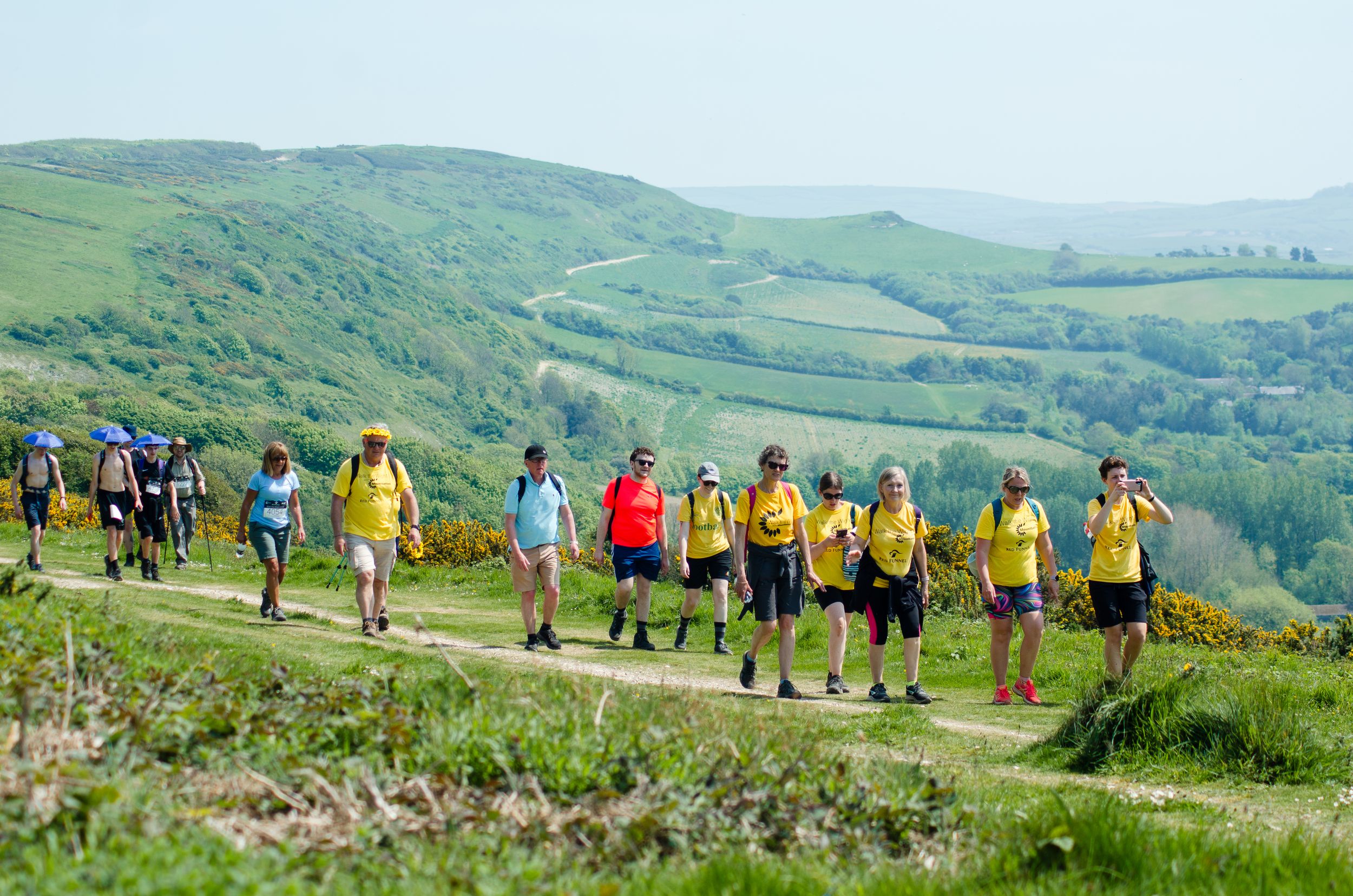 Large group of hikers in colorful attire walking along green hillside trail.