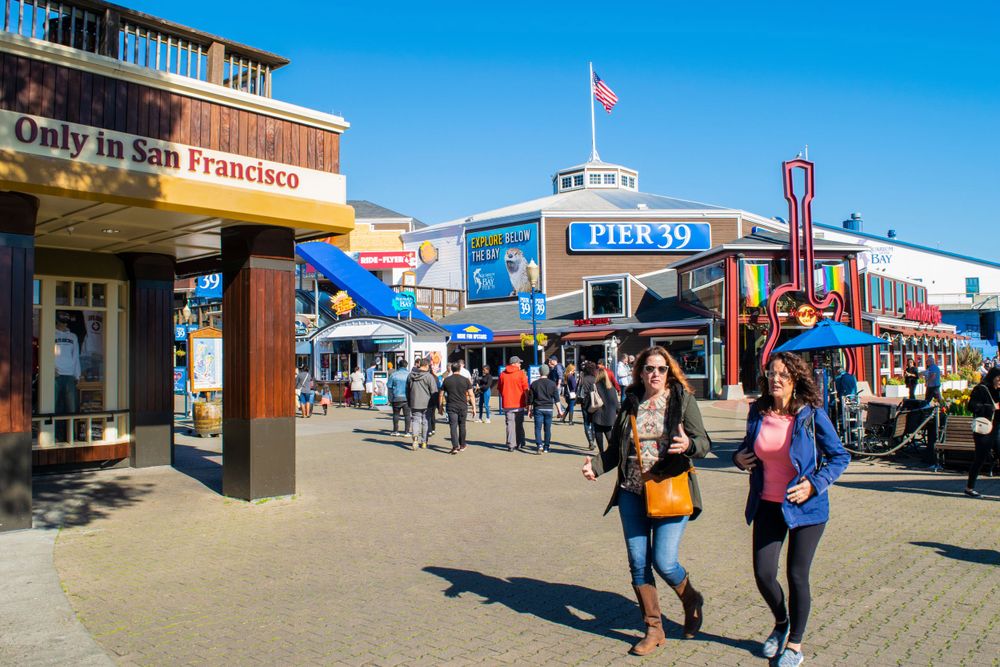 Tourists stroll along Pier 39's bustling waterfront shopping area in San Francisco on a sunny day.