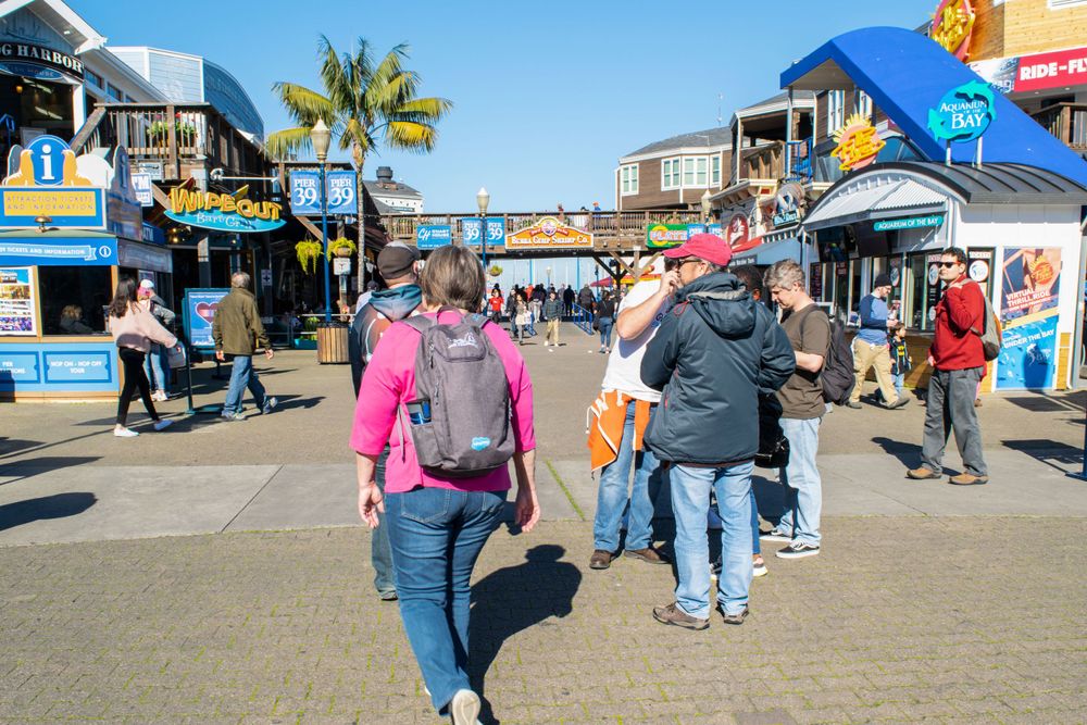 Colorful tourist shops and vendor stalls line a busy waterfront boardwalk on a sunny day.