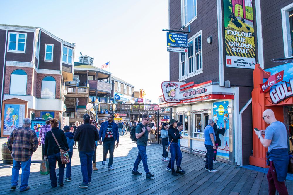 Visitors explore the crowded boardwalk at Pier 39 with shops and restaurants visible along the walkway.