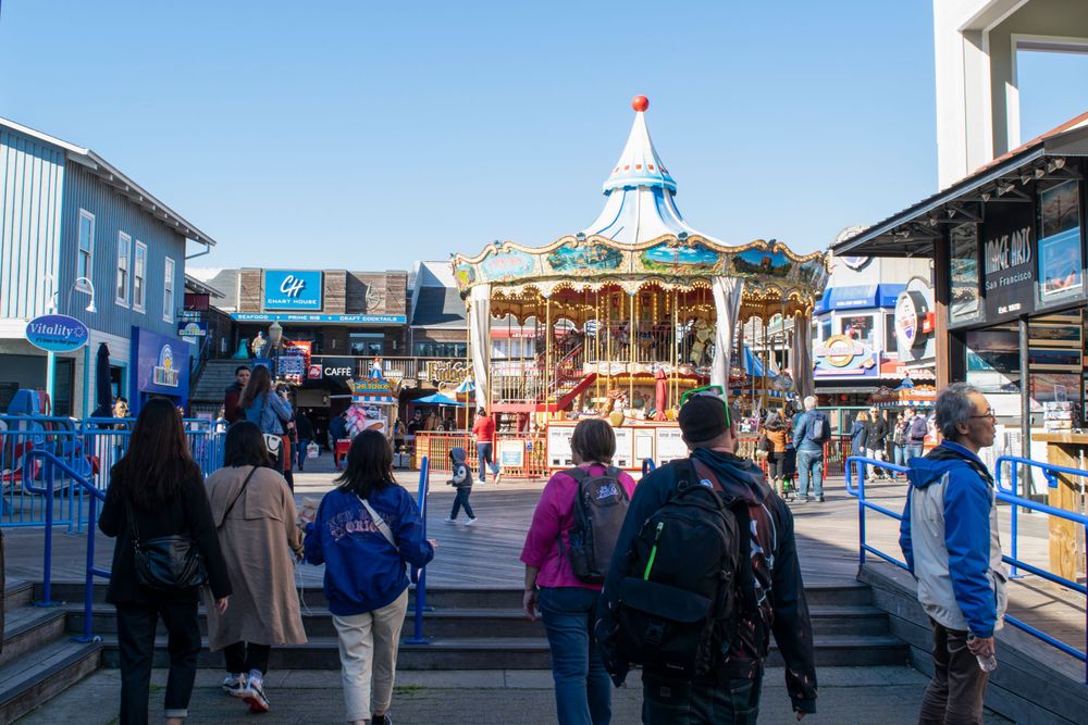 Colorful carousel at Pier 39 surrounded by visitors and blue buildings under clear sky.