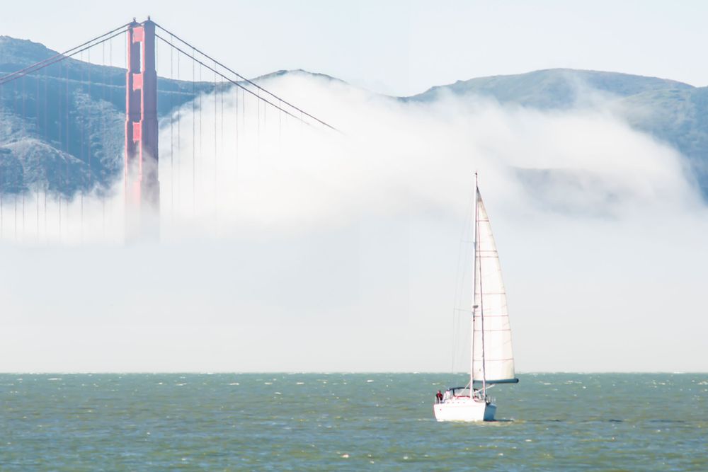 A sailboat cruises past the iconic Golden Gate Bridge shrouded in fog over San Francisco Bay.