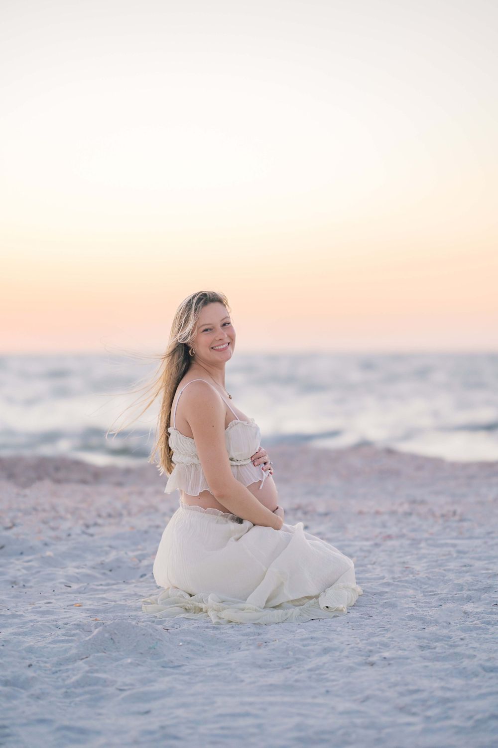 Expectant mother in white dress sitting on beach during sunset.
