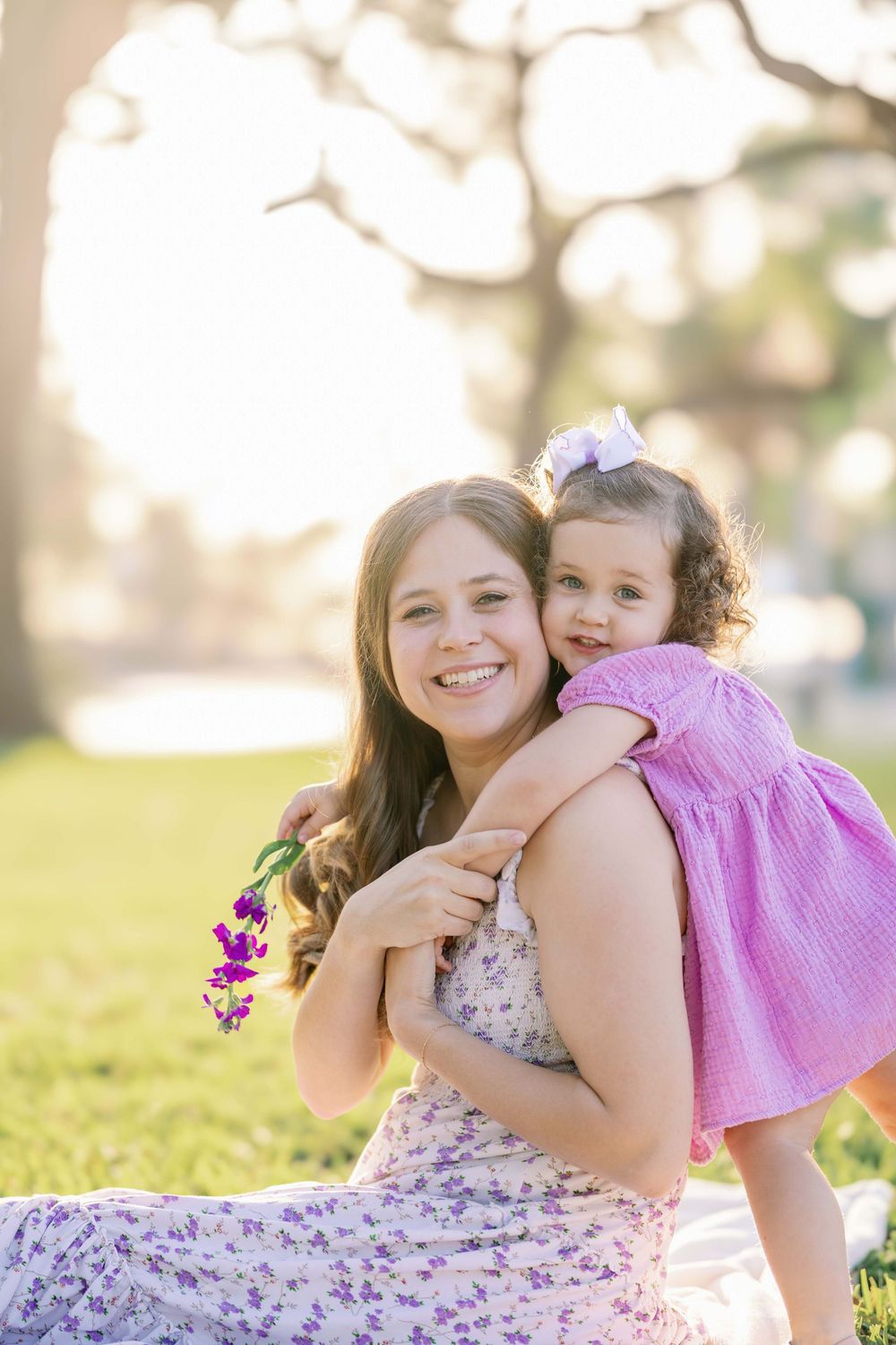 Beautiful outdoor family portrait with two people sitting in grass on sunny day, wearing floral and pink dresses.