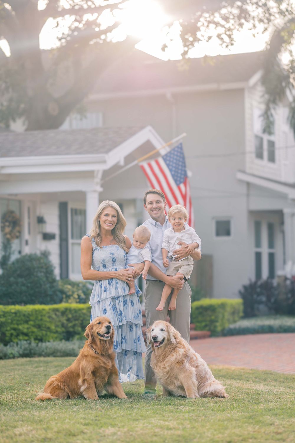 A family with two golden retrievers poses together in front of a house with an American flag.