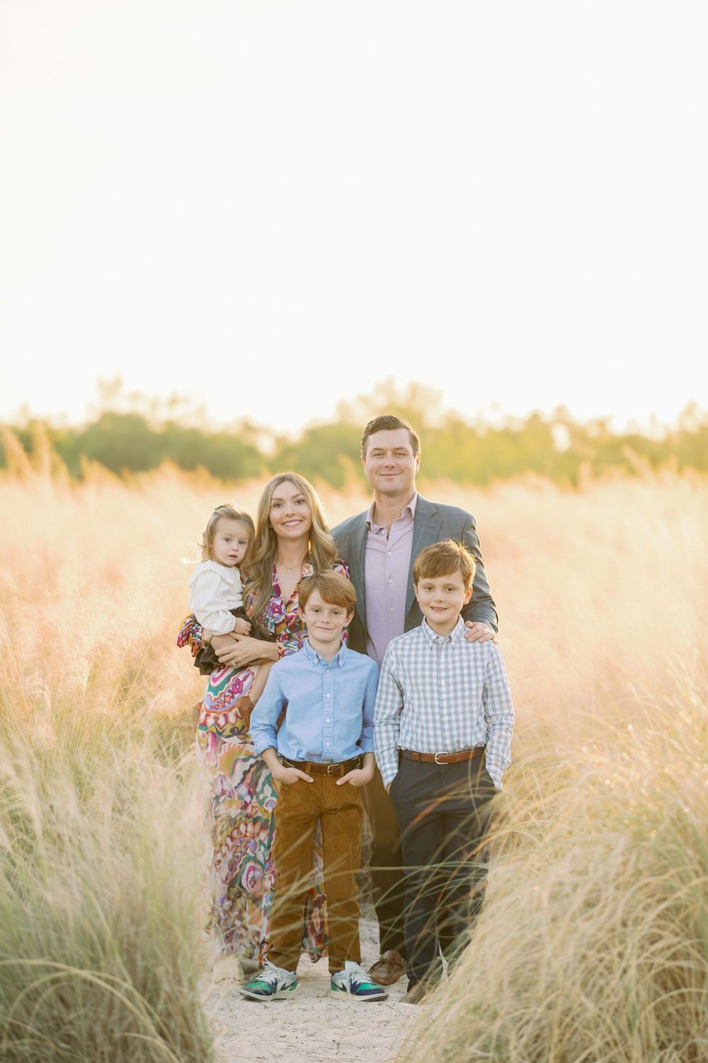 Family of five standing in tall golden grass field during warm sunset outdoor portrait session.