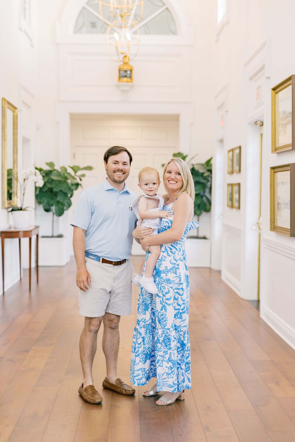 Family portrait in elegant white hallway with gold-framed artwork and hardwood floors.