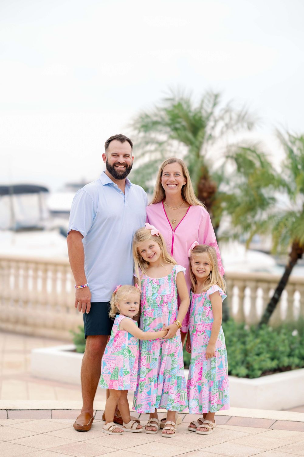 Family of five posing outdoors near a marina with palm trees, wearing coordinated pink and blue outfits.