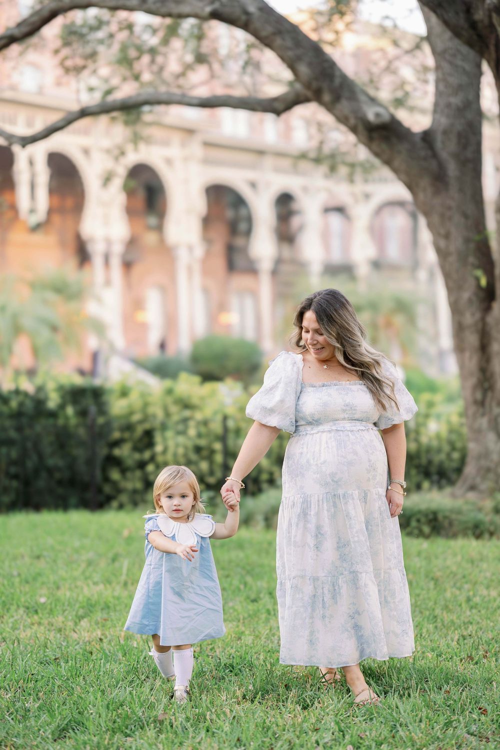 Pregnant mother in light blue dress holds hands with toddler daughter on green lawn near historic building.