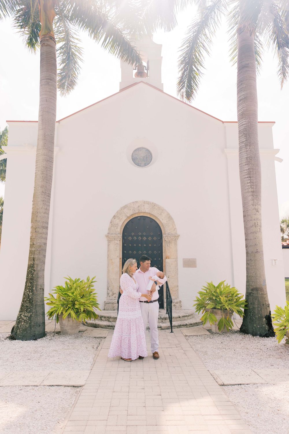 A couple stands with a baby outside a white Spanish-style church with palm trees.