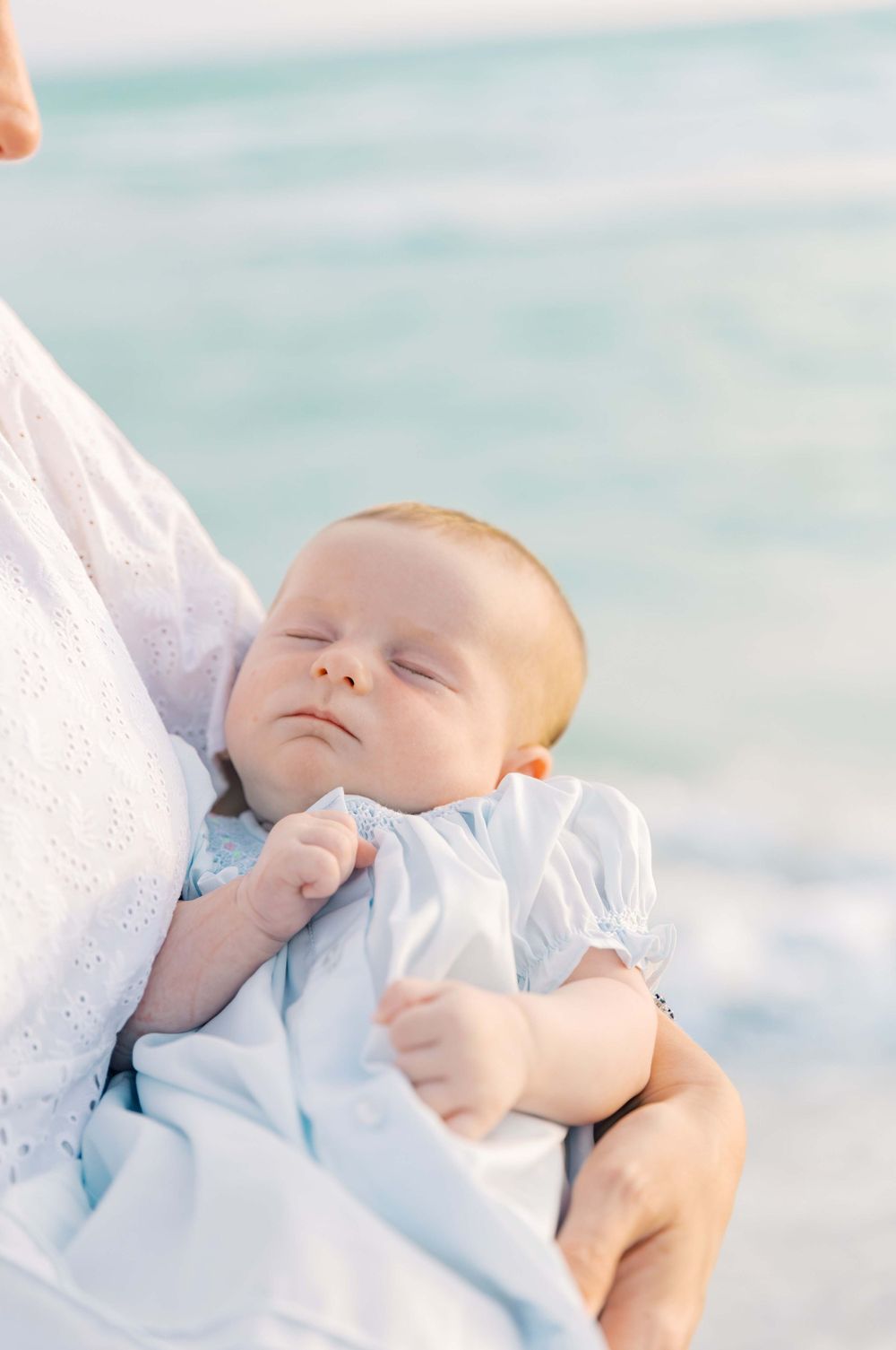 A sleeping baby in light blue clothing rests peacefully against white fabric.