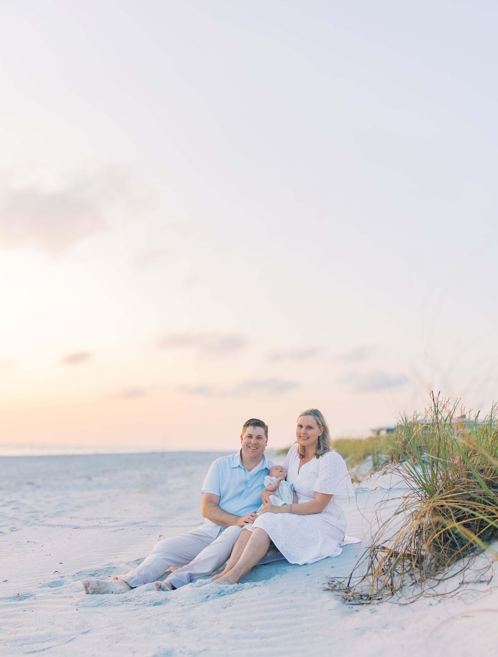 A couple sits together on a sandy beach at sunset with dune grass in the background.