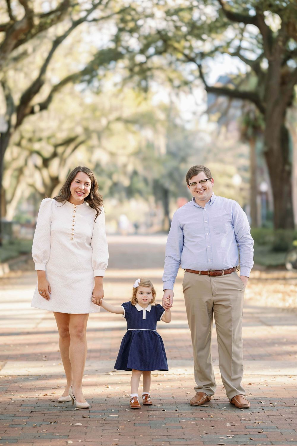 Family of three posing on a tree-lined brick path, parents holding toddler girl in navy dress between them.